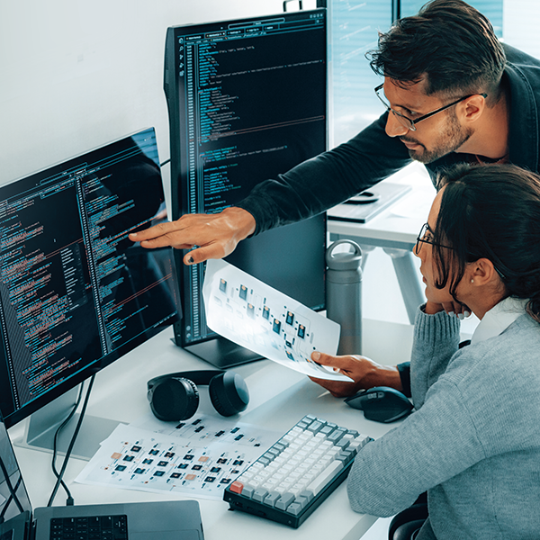 Two people compare data on a piece of paper with information on a computer screen.