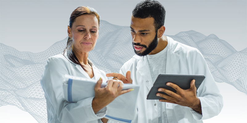 A man and a woman in lab coats stand beside each other, looking at data points on a spreadsheet and tablet. They are standing in front of a grey geometric background. 